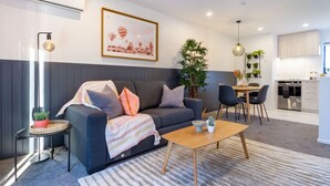 Ein modernes Wohnzimmer mit einem grauen Sofa, einer gestreiften Decke und einem Holztisch. Im Hintergrund ist eine Essecke mit Tisch und Stühlen zu sehen.