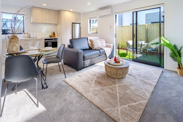 Una sala de estar moderna con un sofá gris, una mesa de comedor con sillas, una zona de cocina y un ventanal corredizo que da a un patio con mobiliario exterior.