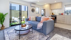 Ein modernes Wohnzimmer mit einem grauen Sofa, einem Glastisch und einem gemusterten Teppich. Eine Glastür führt auf eine Terrasse mit Gartenmöbeln.
