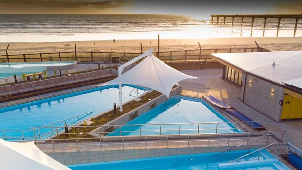 Ein Swimmingpool mit weißem Sonnensegel, Blick auf den Strand und einem Pier im Hintergrund.