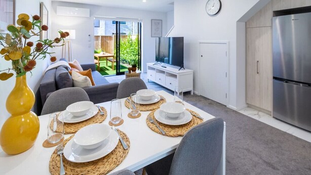 A modern living room with a dining table set for four, a sofa, a TV, and a kitchen area with a refrigerator.