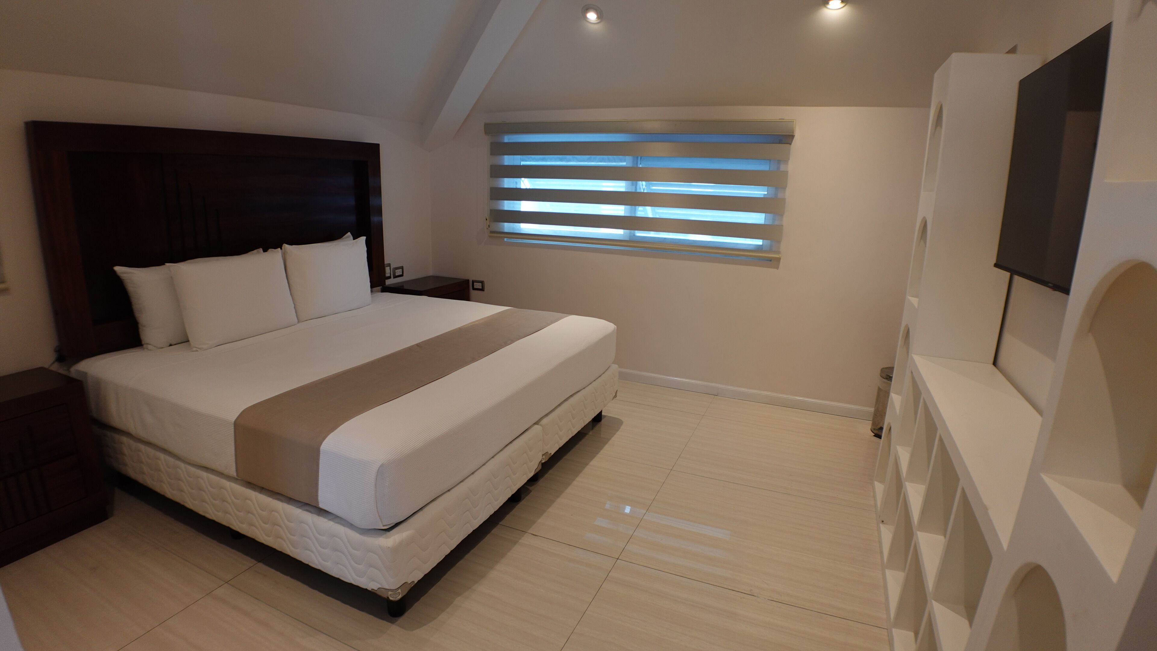 Un dormitorio con cama, mesita de noche, ventana con persianas y televisor montado en la pared.
