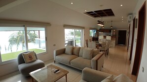 A spacious living area with a sofa, armchairs, and a coffee table. There is a sliding glass door leading to a balcony with a view of palm trees. - Bartolome Oceanfront Suites (Isla Mujeres)