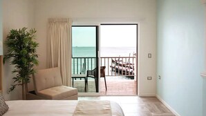 A hotel room with a bed, a chair, a potted plant, and a balcony with a view of the sea and a dock. - Bartolome Oceanfront Suites (Isla Mujeres)
