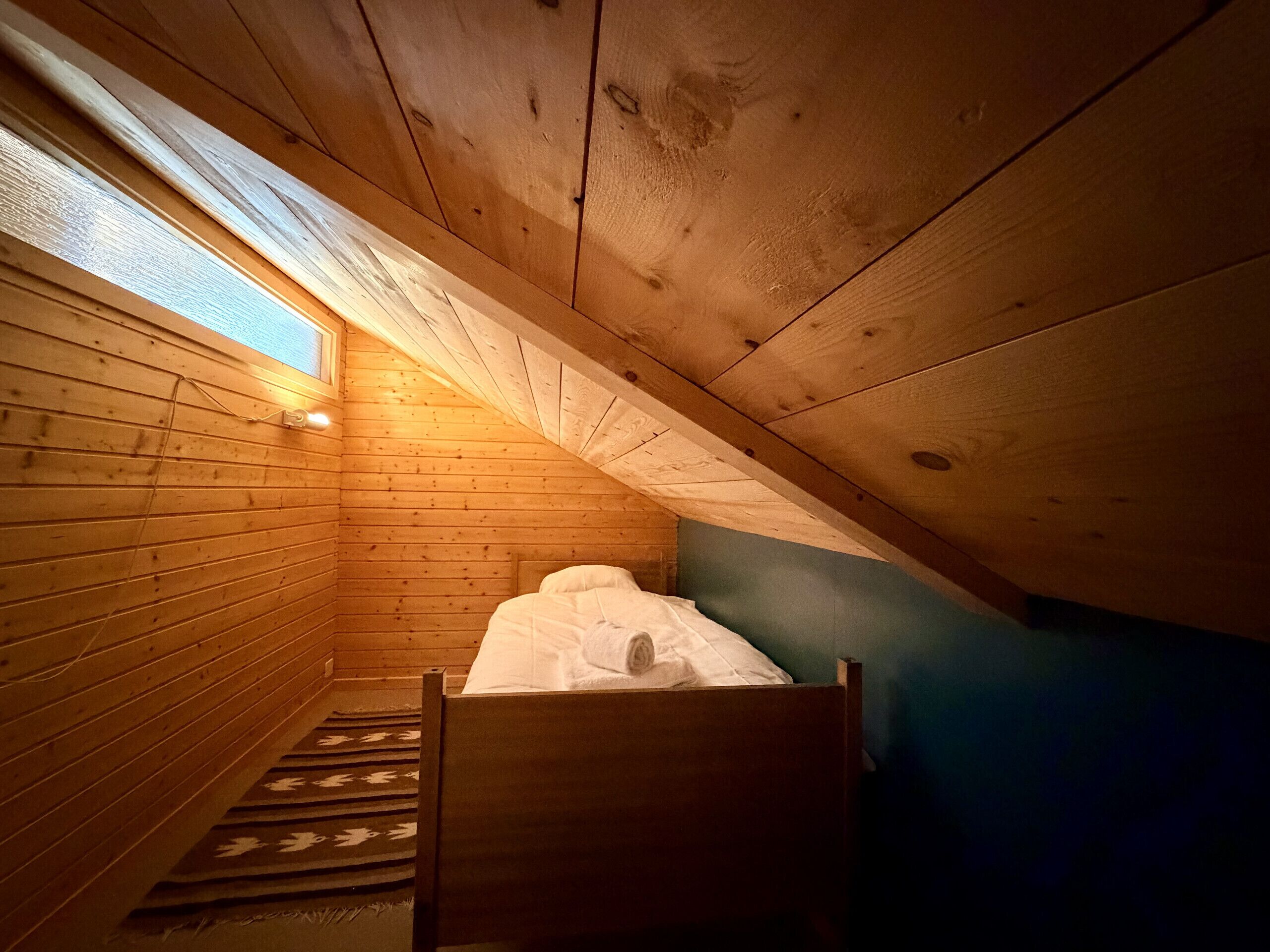 A small, sloped-roofed room with wooden walls, a single bed, and a window.
