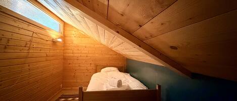 A small, sloped-roofed room with wooden walls, a single bed, and a window.