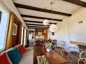 Ein gemütliches Wohnzimmer mit einem Holzespeisetisch, Korbstühlen, einer Couch mit bunten Kissen und einer Küche im Hintergrund.