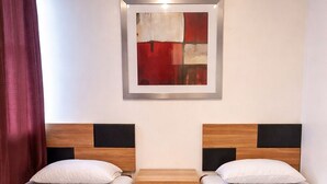 A hotel room with two single beds, a wooden headboard, a small bedside table with a drawer, and a framed abstract painting on the wall.