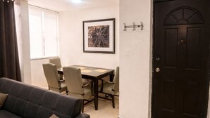A living room with a dark sofa, a dining table with chairs, a framed picture on the wall, and a black door.