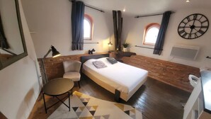 A hotel room with a bed, a desk, a chair, a lamp, and a clock on the wall.