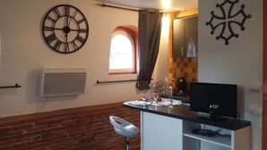 A compact kitchen with a large wall clock, a window with curtains, a small dining area with a table set for two, and a TV on a stand.