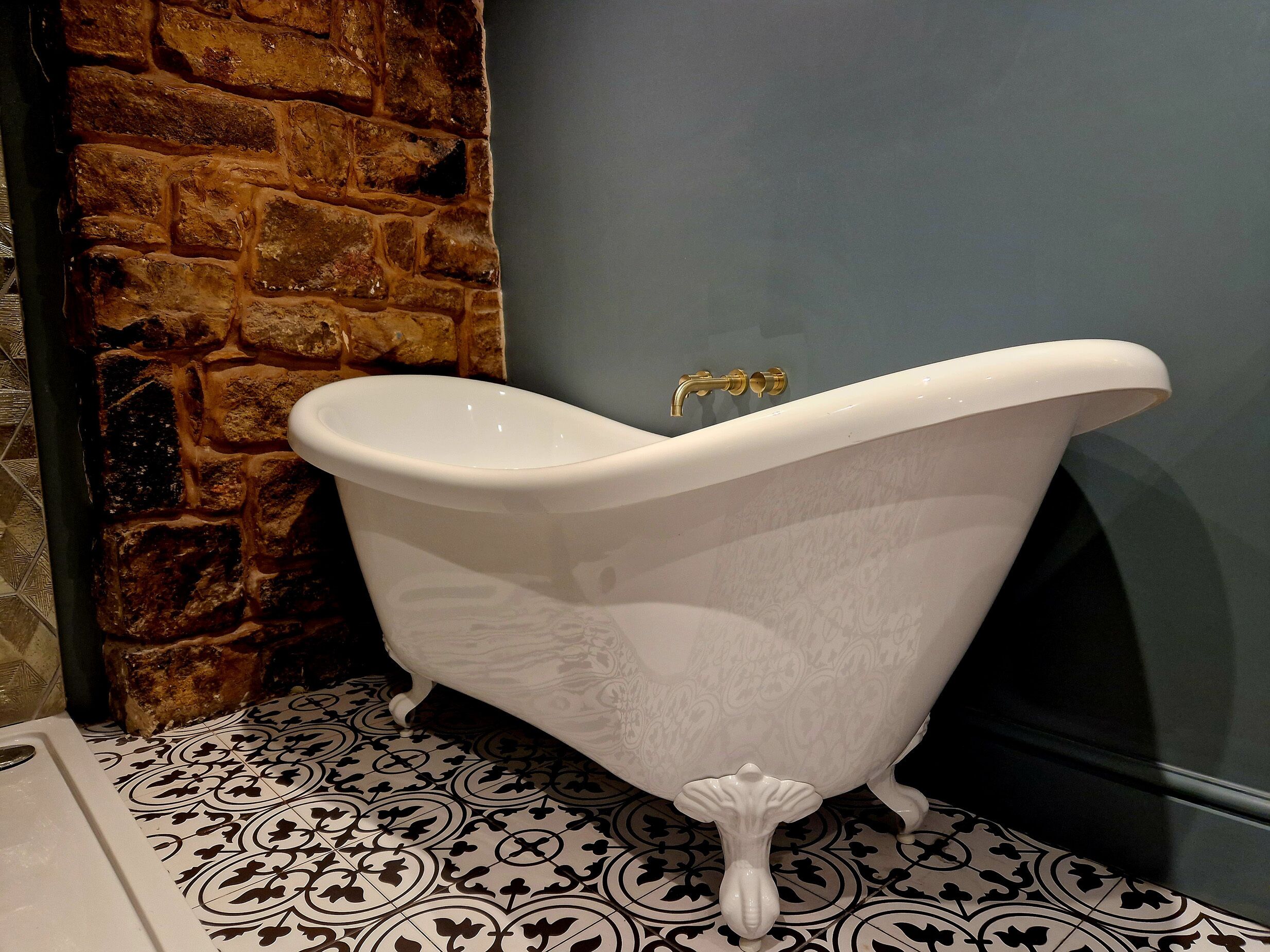 A white clawfoot bathtub with a gold faucet in a bathroom with patterned floor tiles and a stone wall.