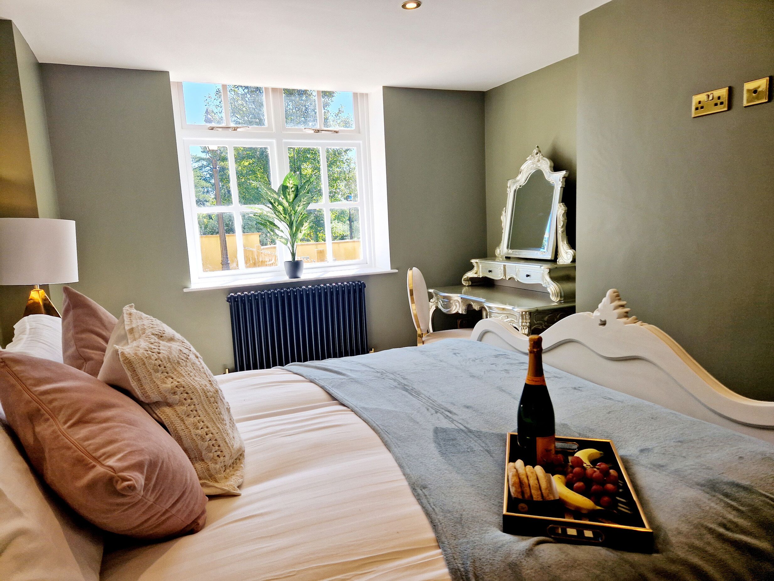 A bedroom with a bed, a bedside table with a bottle and fruit, a dresser, a mirror, and a window with a plant.