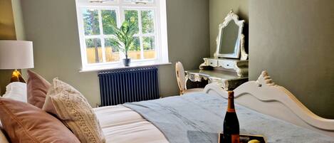 A bedroom with a bed, a bedside table with a bottle and fruit, a dresser, a mirror, and a window with a plant.