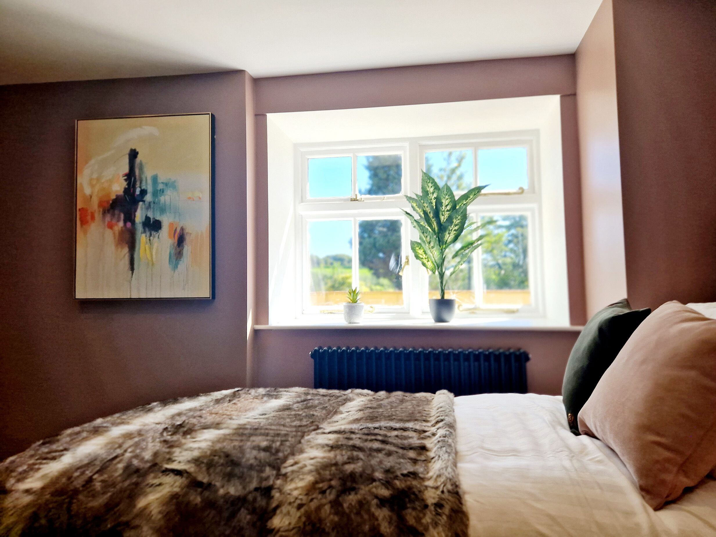 A bedroom with a bed, a window with a view, a radiator, and a painting on the wall.