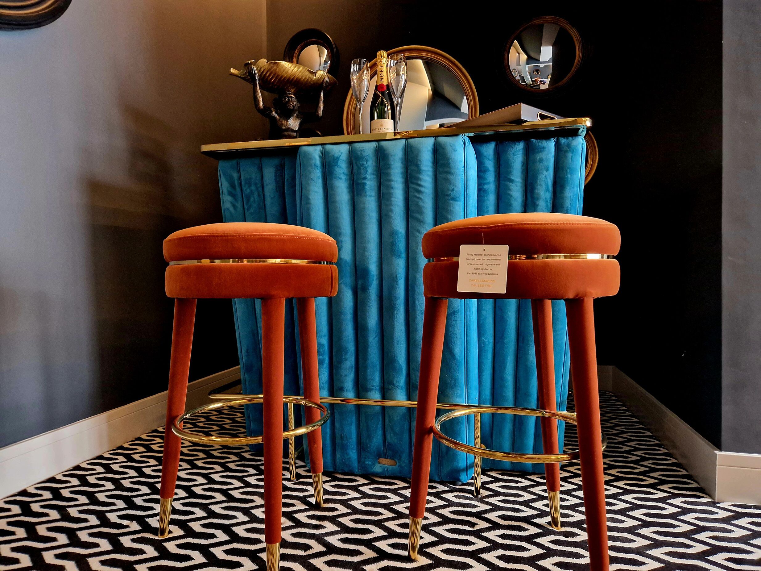 Dos taburetes naranja con patas doradas y una mesa azul tapizada en terciopelo, con un espejo y una botella de champán.