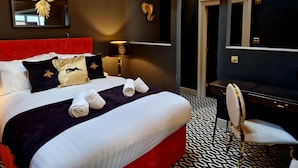 Habitación de hotel con cabecera tapizada en rojo, una cama grande con ropa de cama blanca y negra, un adorno dorado sobre la cama, un escritorio con silla y un suelo con estampado.