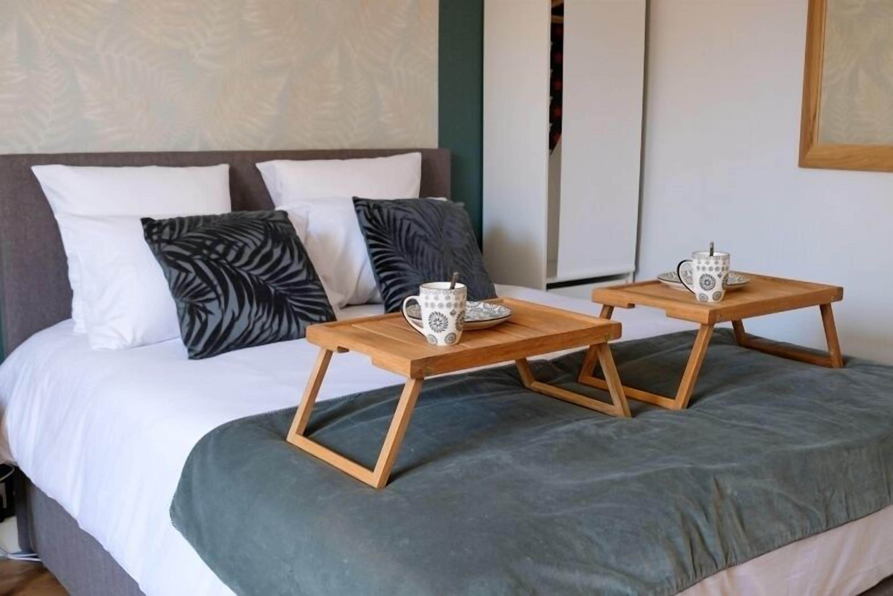Una cama bien tendida con un edredón gris y dos mesitas de noche de madera, cada una sosteniendo una taza blanca con diseño negro.