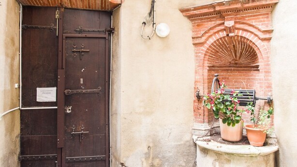 A rustic building with a large wooden door and a small arched window with a water spigot.