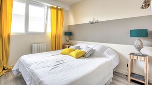 Una cama bien tendida con ropa blanca y dos almohadas amarillas, una mesita de noche con lámpara y una ventana con cortinas amarillas.