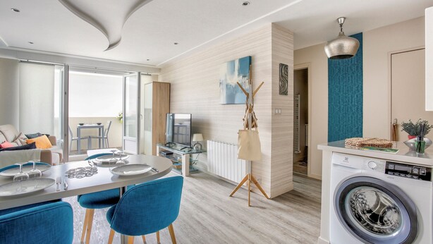 Una cocina moderna con lavadora integrada, un comedor con sillas azules y un amplio ventanal con vistas al exterior.