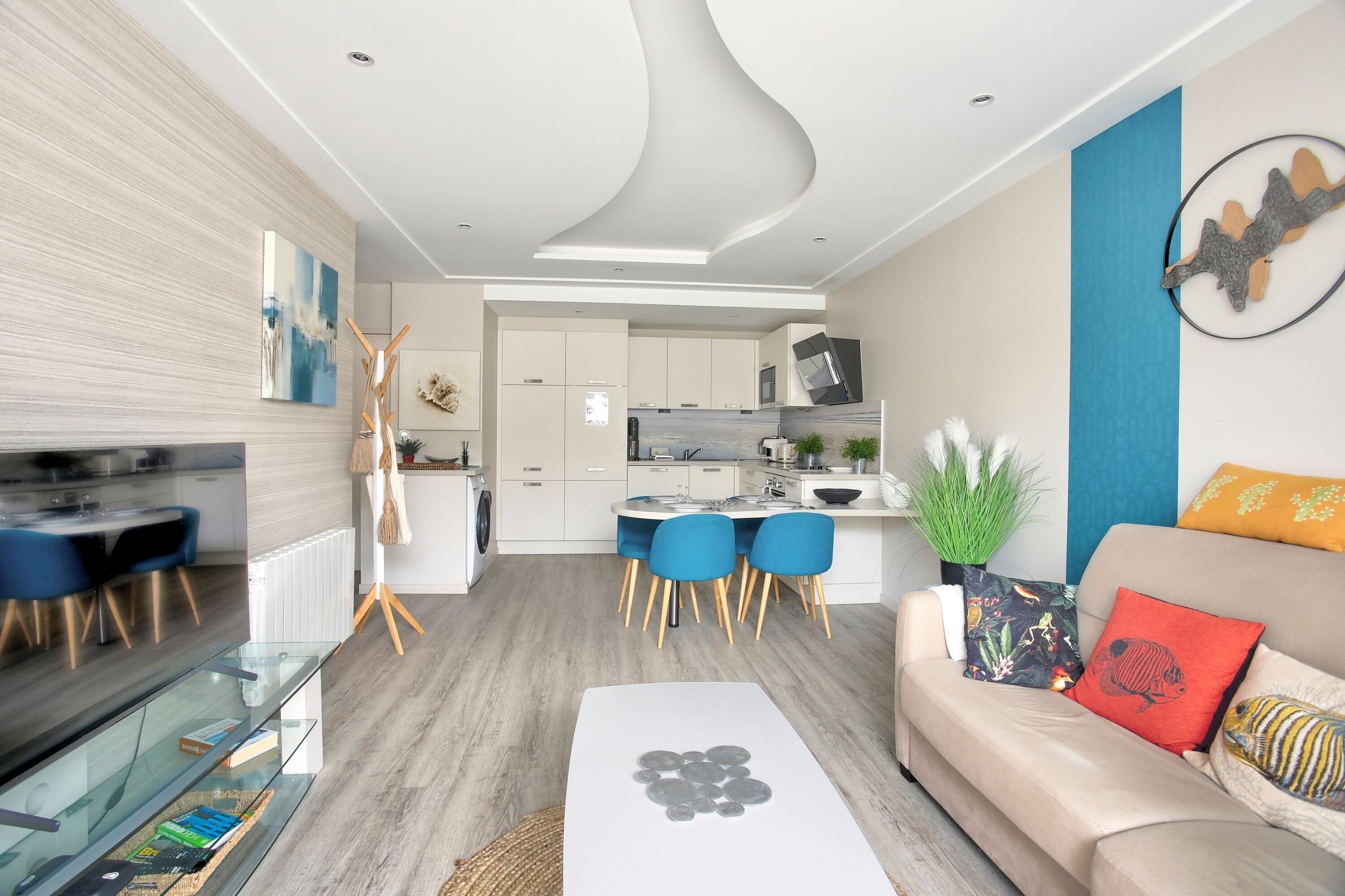 En moderne stue med et glasbord, en beige sofa med farverige puder, samt et spiseområde med blå stole og et hvidt bord.