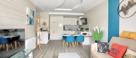 En moderne stue med et glasbord, en beige sofa med farverige puder, samt et spiseområde med blå stole og et hvidt bord.