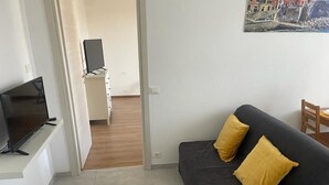 Ein Wohnzimmer mit einem schwarzen Sofa, zwei gelben Kissen, einem Flachbildfernseher auf einem Standfuß und einem Gemälde an der Wand.