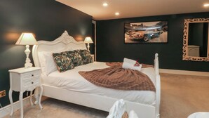 A bedroom with a white bed, two bedside tables with lamps, a dark wall with a car picture, and a mirror with an ornate frame. - Alexander (Preston)