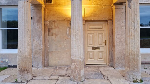 A building entrance with a wooden door, flanked by stone columns, and a small window to the left. - Alexander (Preston)