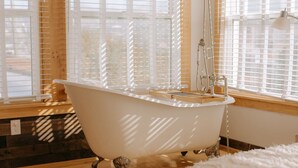 Une baignoire blanche, sur pieds, se trouve dans une pièce dont le sol est recouvert de parquet et qui possède de grandes fenêtres équipées de stores.