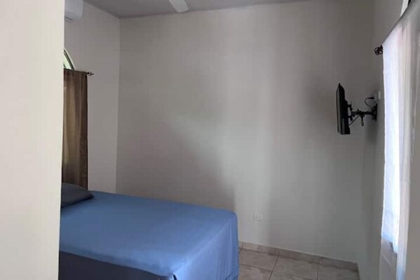 Une petite chambre avec un lit simple, une couverture bleue, un téléviseur fixé au mur et un rideau sur le côté gauche.