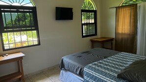 Ein Schlafzimmer mit einem Bett, einem kleinen Holztisch, einem an der Wand montierten Fernseher und zwei Fenstern mit Blick auf Grünflächen.