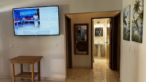 Ein Flur mit einem Fernseher, der eine Nachrichtensendung zeigt, einem Holztisch und einem Badezimmer mit Waschbecken und Spiegel.
