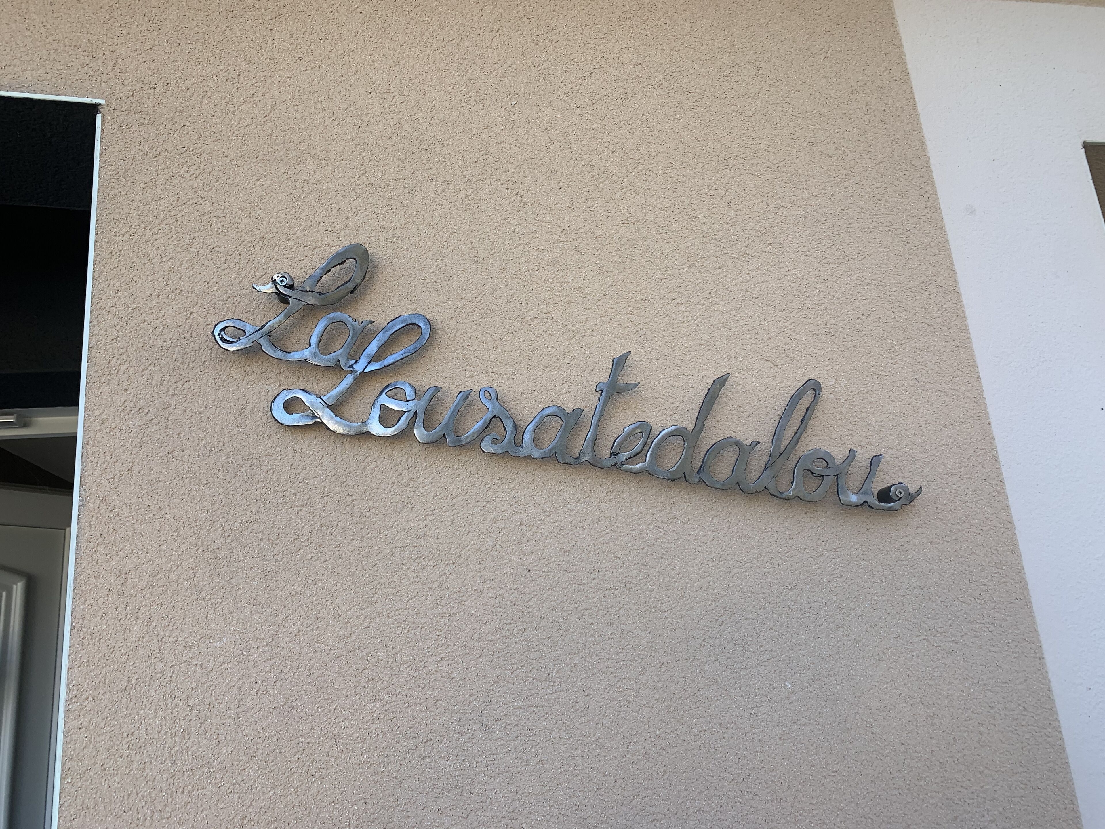 Una señal metálica con el nombre "Lourdesdalu" sobre una pared color beige.