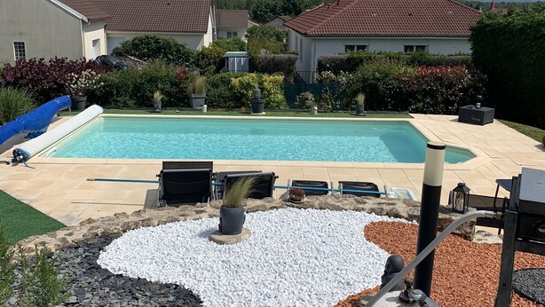 A swimming pool with a pebble design, surrounded by a garden with various plants and a barbecue area.