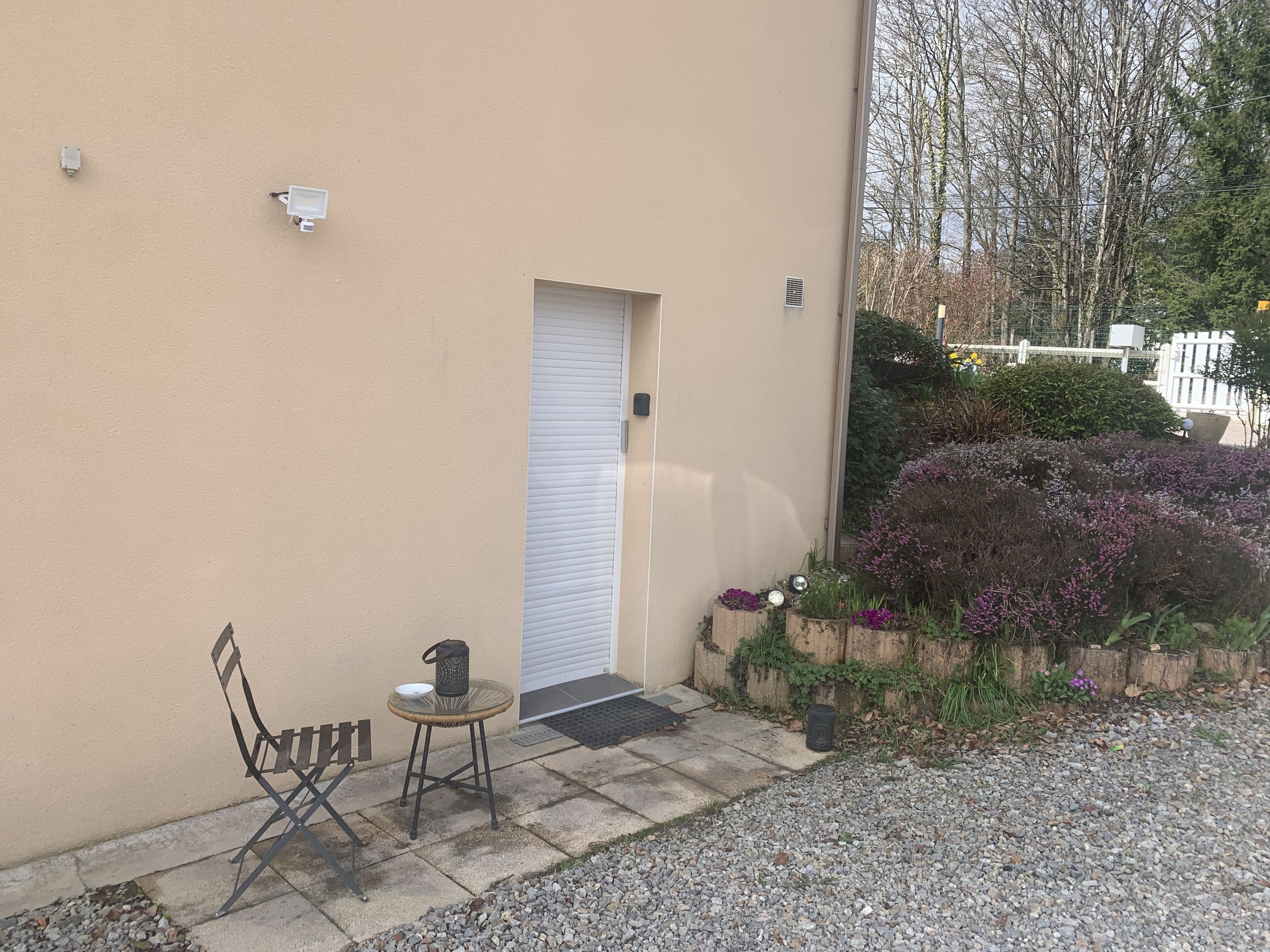 Een gebouw met een beige gevel, een gesloten witte deur, een klein terras met een stoel en tafel, en een tuin met paarse bloemen.