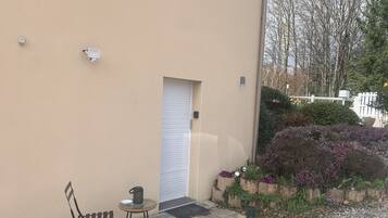 Een gebouw met een beige gevel, een gesloten witte deur, een klein terras met een stoel en tafel, en een tuin met paarse bloemen.