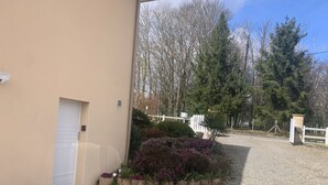 A building with a beige exterior, a closed white door, a small outdoor seating area with a table and chairs, and a gravel driveway leading to a white fence and trees.
