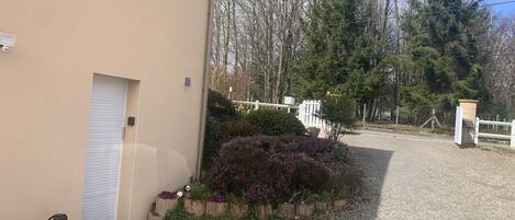 Un edificio con fachada color beige, una puerta blanca cerrada, un pequeño espacio al aire libre con mesa y sillas, y un camino de grava que conduce a una cerca blanca y árboles.