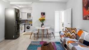 Une cuisine moderne avec des armoires blanches, un réfrigérateur en acier inoxydable et un lave-linge. On y trouve un coin repas avec une table et des chaises blanches, ainsi qu’un tapis coloré sur le sol.