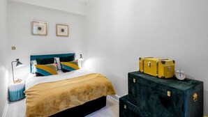 Une chambre à coucher avec un lit, deux tableaux encadrés au mur, un lustre et deux valises posées sur une commode.