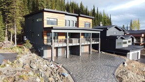A modern two-story house with a balcony, surrounded by a rocky landscape and dense forest. - Cozy duplex on ski way with private hot tub, laundry, garage and large ski room! (Silver Star Mountain)