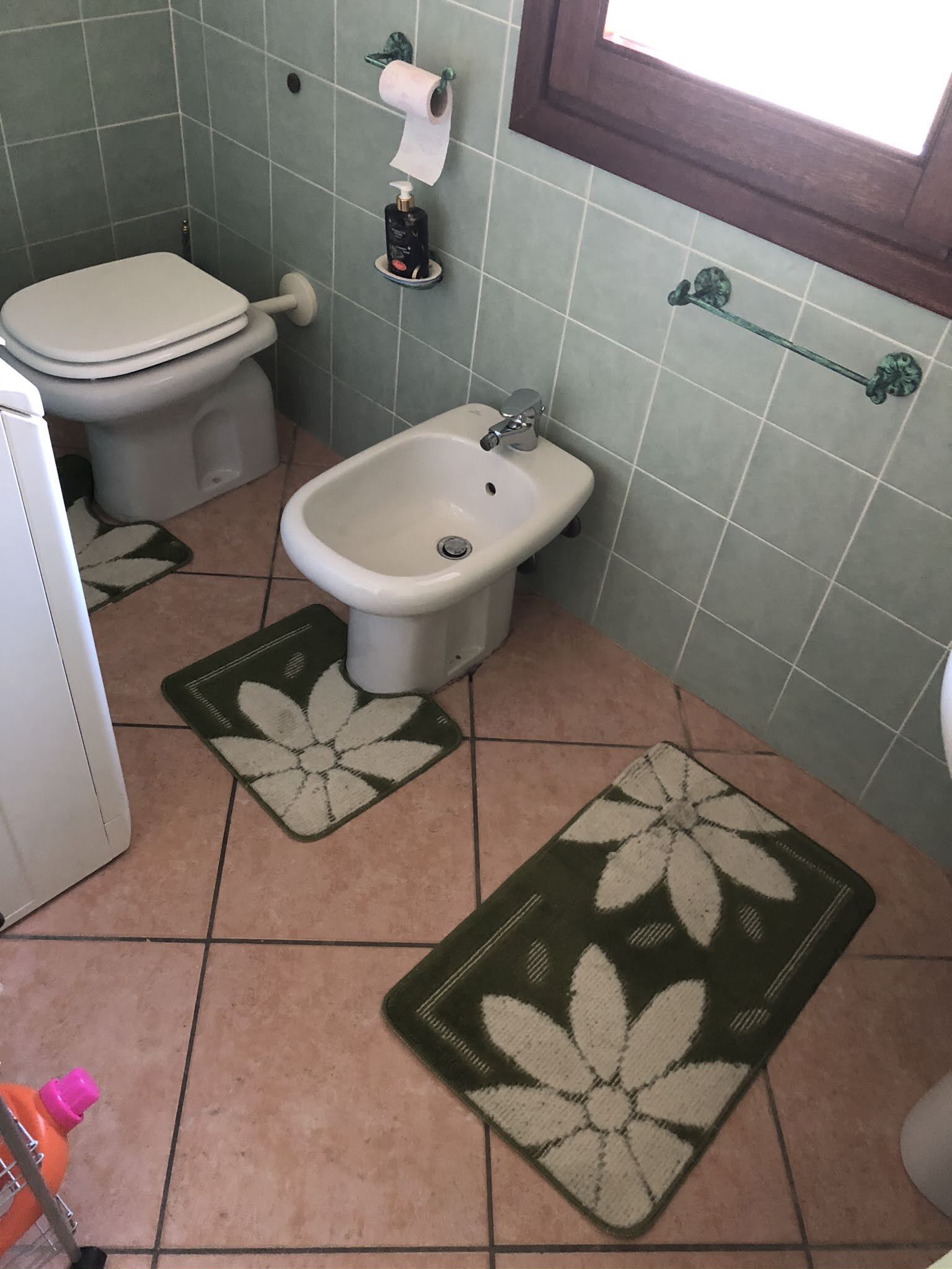 Ein Badezimmer mit Toilette, Bidet und Waschbecken. Auf dem Boden liegen zwei Teppichmatten mit Blumenmuster.