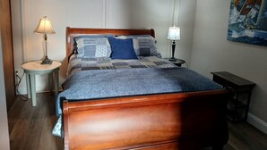 A neatly arranged bedroom with a wooden bed, two bedside tables with lamps, a large window, and a painting on the wall. - Cottage/Studio in Cobble Hill Village - hiking, wineries and more (Cobble Hill)