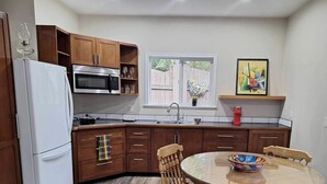 A compact kitchen with wooden cabinets, a refrigerator, a microwave, and a dining area with a round table and chairs. - Cottage/Studio in Cobble Hill Village - hiking, wineries and more (Cobble Hill)