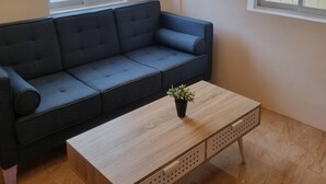 Een moderne woonkamer met een donkerblauwe bank, een houten salontafel met een plant, en grote ramen met zonwering.