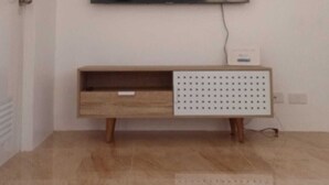 Een kamer met een wandgemonteerde flatscreen-tv aan een witte muur, een houten bank met een geperforeerd paneel en een betegelde vloer.