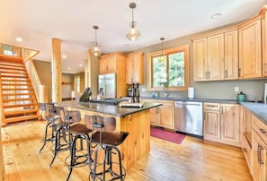 A spacious kitchen with wooden cabinets, a central island with bar stools, stainless steel appliances, and a staircase leading to another level.