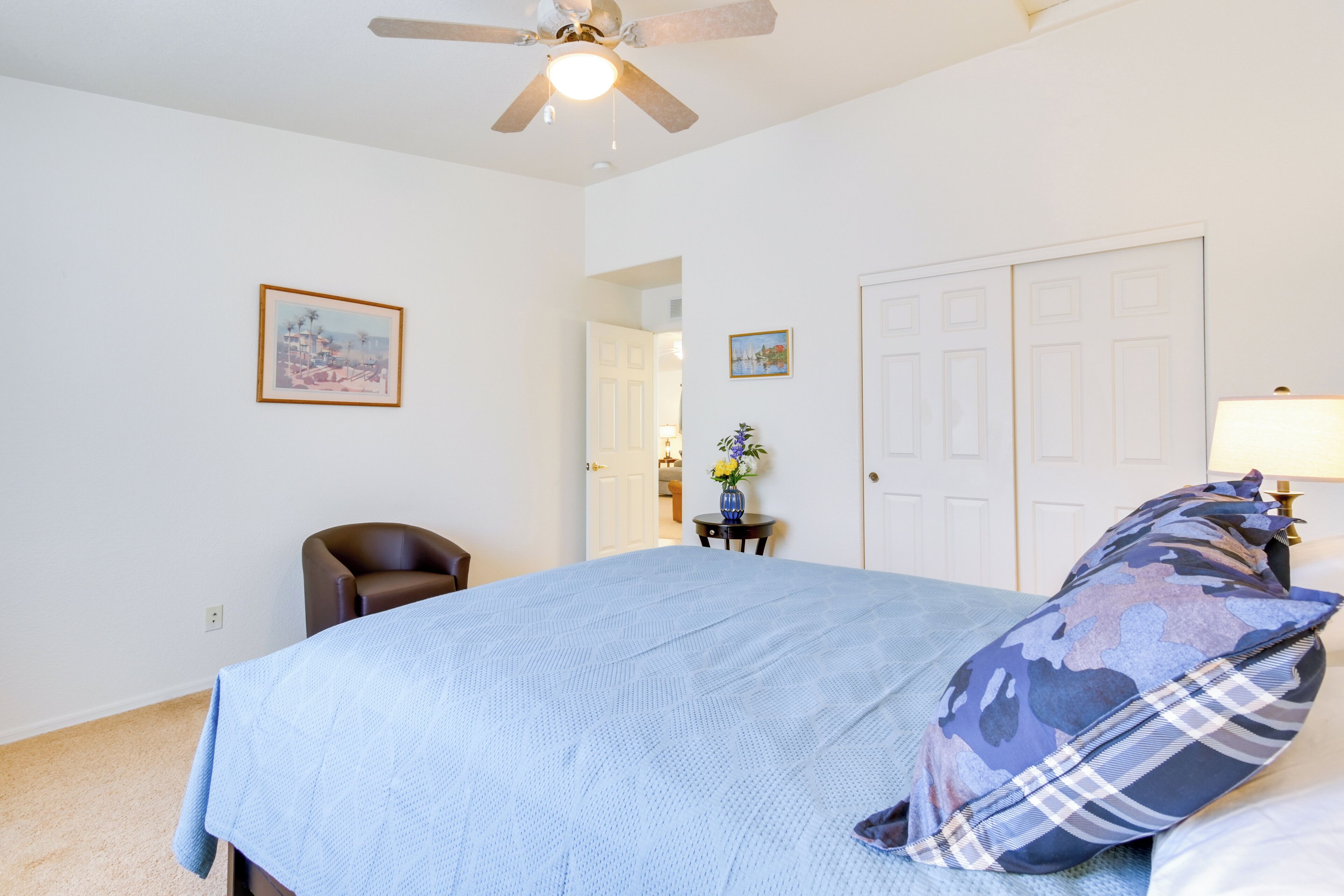 A bedroom with a bed, a ceiling fan, a chair, a desk with a vase, and a door.